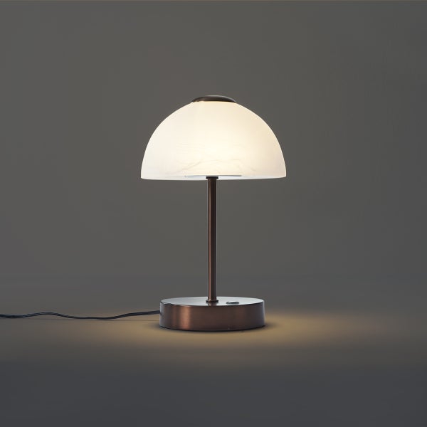 Tischleuchte mit bronzefarbenem Metallfuß und kuppelförmigem Schirm aus Glas in Alabaster-Optik, eingeschaltet.