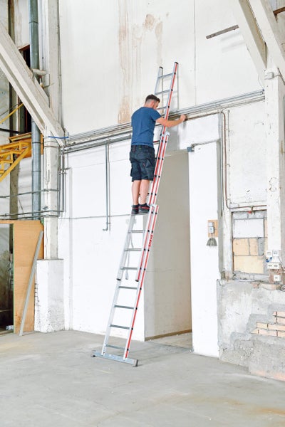 Ein Mann steht auf einer Mehrzweckleiter und führt Arbeiten an einer Wand in einem Innenraum durch.