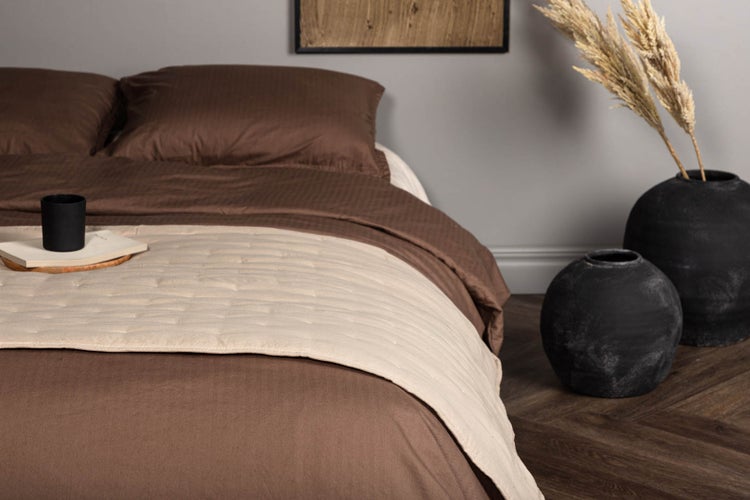 Schlafzimmer mit brauner Bettwäsche, beige gesteppter Tagesdecke und dunklem Fischgrätenparkett mit dekorativen Vasen.