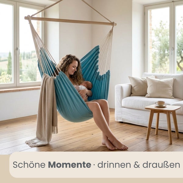 Heller Wohnraum mit Parkettboden, in dem eine Frau mit einem Baby in einem blauen Hängesessel aus Stoff sitzt. Im Hintergrund ein weißes Sofa.