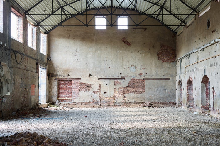 Innenansicht einer leerstehenden Fabrikhalle mit rohen Backsteinmauern, sichtbarer Stahlkonstruktion und einem Boden aus Schotter.