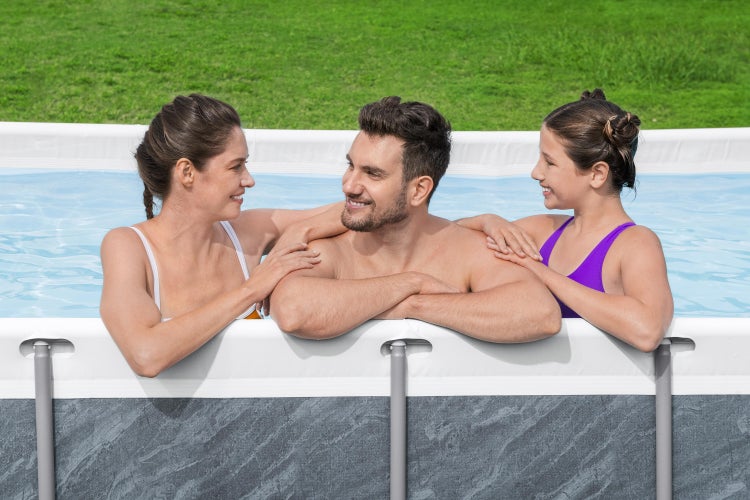 Familie in einem rechteckigen Aufstellpool mit grauer Steinoptik im Garten auf einer Rasenfläche.