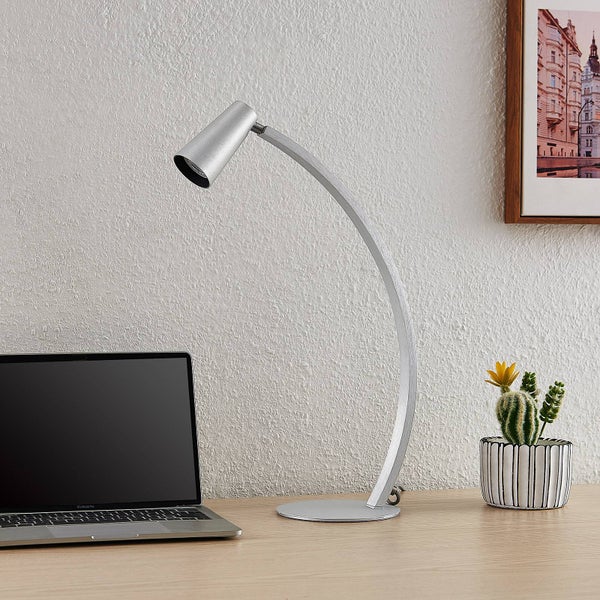 Moderne silberne Schreibtischlampe mit gebogenem Arm auf einem Holztisch neben einem Laptop und einem kleinen Kaktus.