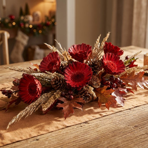 Herbstliches Gesteck mit roten Blumen, Tannenzapfen, Beeren und trockenem Gras auf einem Holztisch als Tischdekoration.