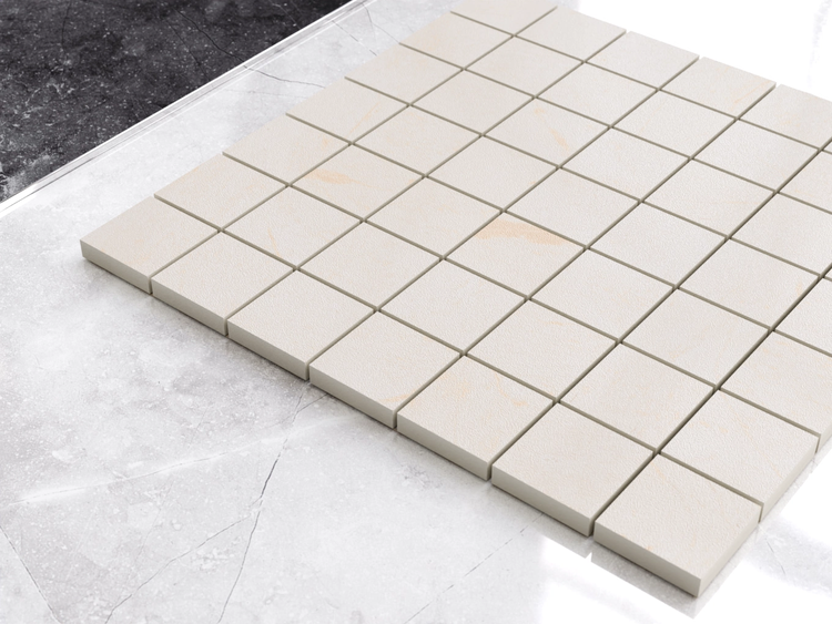 Quadratische Mosaikfliesen in Steinoptik auf einer großen Bodenfliese mit einer Abschlussschiene aus Metall.