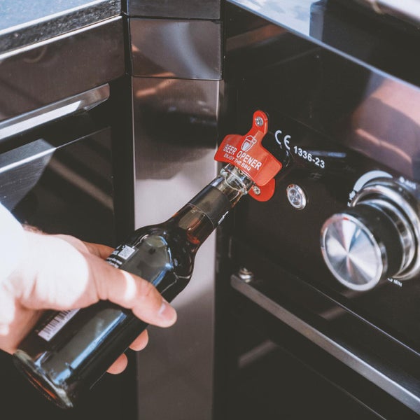 Roter Wandflaschenöffner an einem schwarzen Metallgrill montiert. Eine Hand öffnet eine dunkle Glasflasche am Öffner.