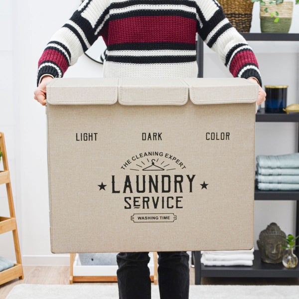 Beige Wäschetruhe mit drei Fächern beschriftet mit Light, Dark und Color, gehalten von einer Person in einem hellen Raum.