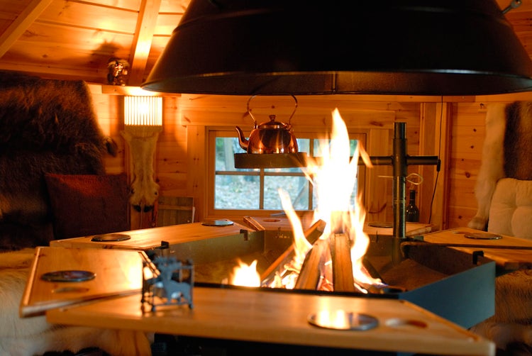 Innenraum einer Kota mit Feuerstelle, Holztischen, Fellen und einem Kupferkessel