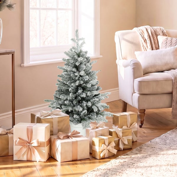 Ein künstlicher, verschneiter Weihnachtsbaum steht in einem Wohnzimmer, umgeben von festlich verpackten Geschenken neben einem Sessel am Fenster.