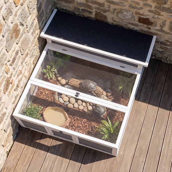 Schildkrötengehege aus Holz mit Gitterabdeckung und klappbarem Dach, eingerichtet mit Rindenmulch, Steinen und Pflanzen auf einer Holzterrasse.
