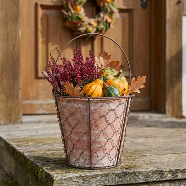 Tontopf mit Drahtkorb und herbstlicher Bepflanzung mit Heidekraut und Zierkürbissen vor einer Holztür als Herbstdekoration.