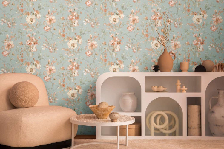 Vliestapete mit floralem Blumenmuster an einer Wohnzimmerwand kombiniert mit einem beigen Sessel und einem weißen Regal mit Dekoration.
