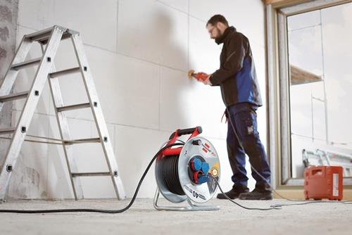 Bauarbeiter bei der Arbeit mit Kabeltrommel, Leiter und Schleifwerkzeug an einer Wand