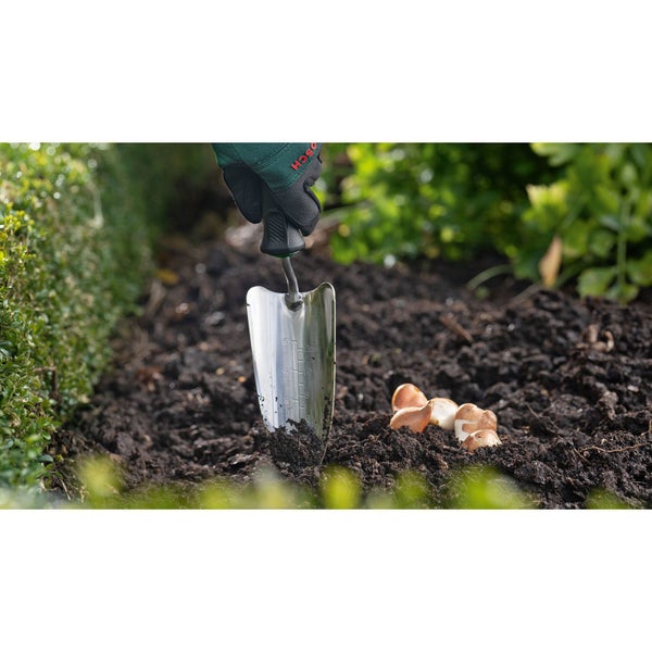 Szene der Gartenarbeit mit einer Hand, die mit einer rostfreien Pflanzkelle Blumenzwiebeln in die Erde setzt.