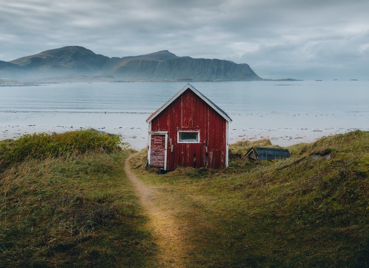 Rotes Holzhaus an einer Küste mit Bergen im Hintergrund, Dünenlandschaft und Sandstrand unter bewölktem Himmel.