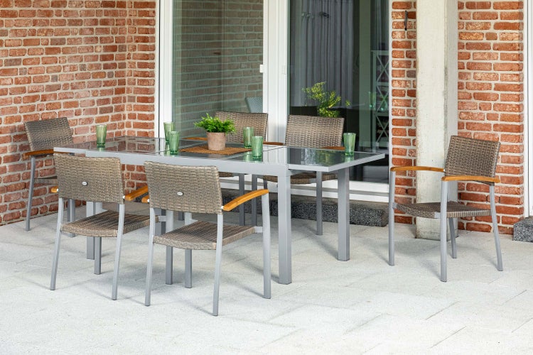 Gartenset bestehend aus einem grauen Aluminiumtisch mit Glasplatte und sechs Flechtstühlen mit Holzarmlehnen auf einer gepflasterten Terrasse.