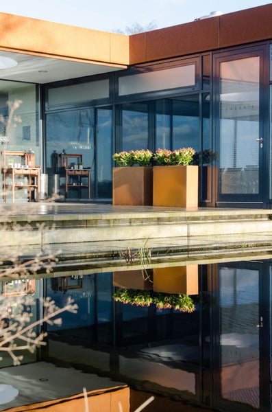 Zwei quadratische Pflanzgefäße in Rostoptik auf einer modernen Terrasse vor einer Glasfassade, die sich in einem Teich spiegeln.