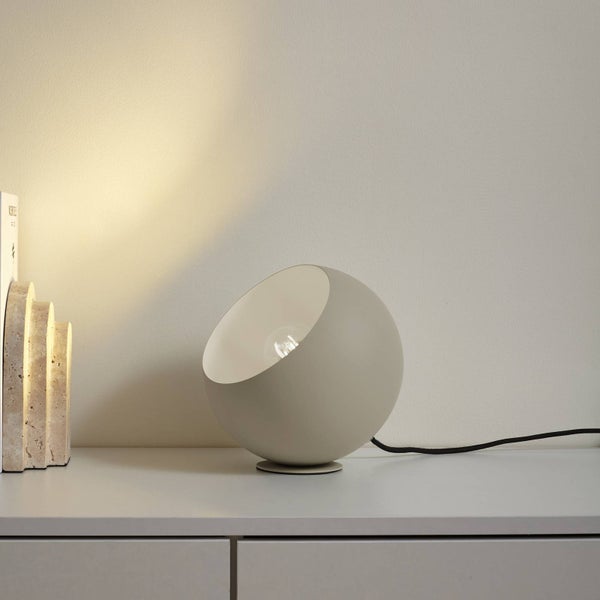 Kugelförmige Tischlampe im modernen Design, beige, Metall, schräge Öffnung mit sichtbarem Leuchtmittel, auf einer weißen Kommode neben Buchstützen.