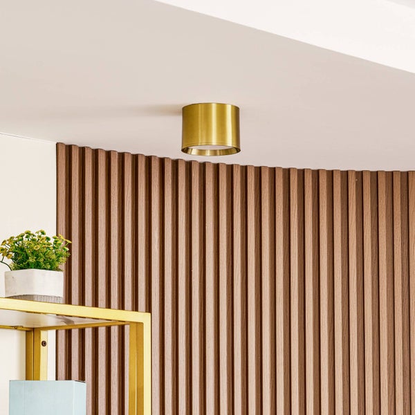 Goldfarbene zylindrische Deckenleuchte vor einer Wand mit Holzlamellen.