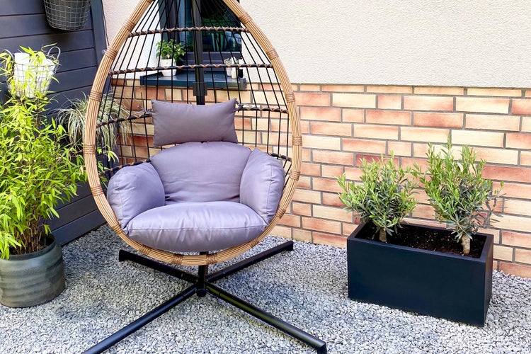 Moderner Hängesessel in Eiform mit Metallgestell, Polstern und Geflecht auf einer Terrasse mit Pflanzen und Kiesboden.