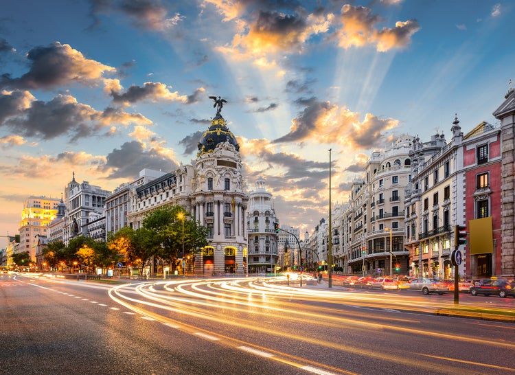 Stadtansicht von Madrid mit dem Metropolis-Gebäude bei Sonnenuntergang, beleuchtete historische Architektur und Lichtspuren von Fahrzeugen.