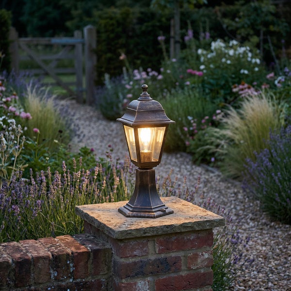 Beleuchtete Gartenlaterne im klassischen Stil auf einem gemauerten Sockel in einem Garten mit Blumenbeeten und Kiesweg.