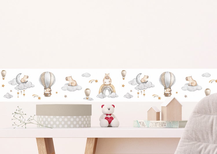 Wandbordüre mit Tiermotiven, Heißluftballons und Wolken in einem hellen Kinderzimmer mit Gitterbett, Rattansesseln und Holzspielzeug.