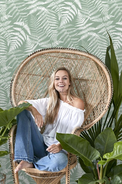 Frau sitzt in einem Rattansessel vor einer Wand mit Farnmuster