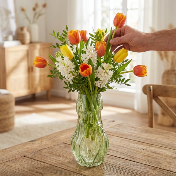 Bunter Frühlingsstrauß mit Tulpen und Hyazinthen in einer strukturierten Glasvase auf einem Holztisch, eine Hand richtet eine Tulpe aus.