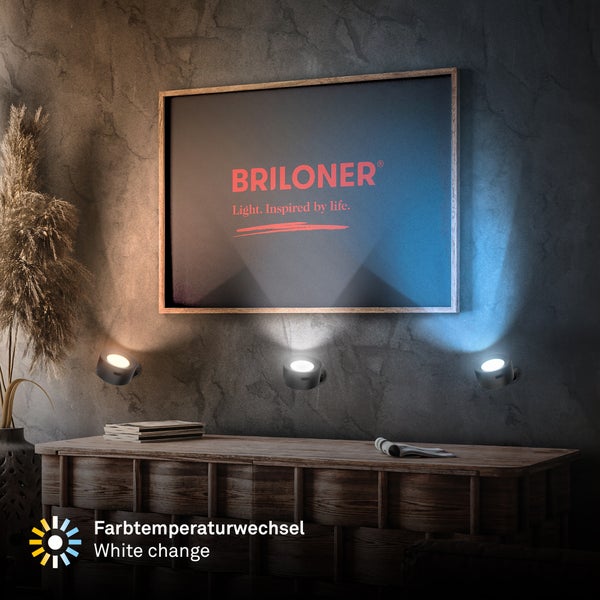 LED-Wandspots mit Farbtemperaturwechsel von warmweiß bis kaltweiß auf einer strukturierten Wand über einem hölzernen Sideboard. Briloner Logo.