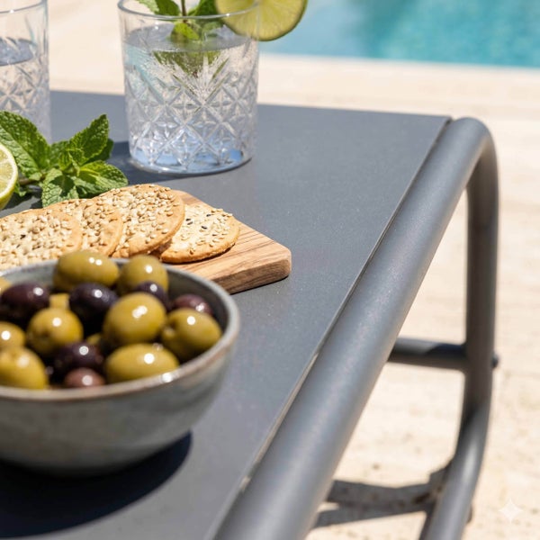 Grauer Gartentisch mit abgerundetem Aluminiumrahmen, darauf Schalen mit Oliven, Crackern und Erfrischungsgetränken am Pool.