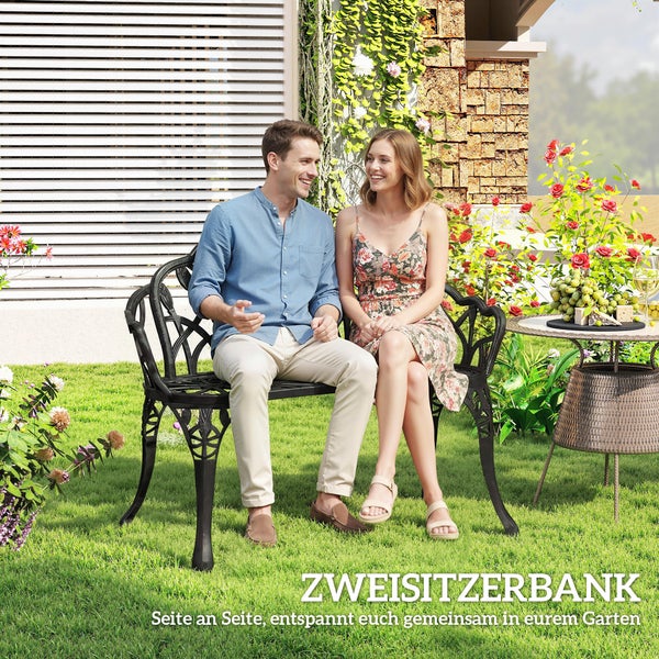 Ein Paar sitzt auf einer verzierten Gartenbank aus Metall in einem hellen Garten neben einem Tisch mit Weintrauben.