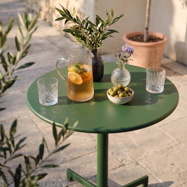 Grüner runder Bistrotisch aus Metall auf einer Terrasse mit Krug, Gläsern und Oliven in heller Atmosphäre.
