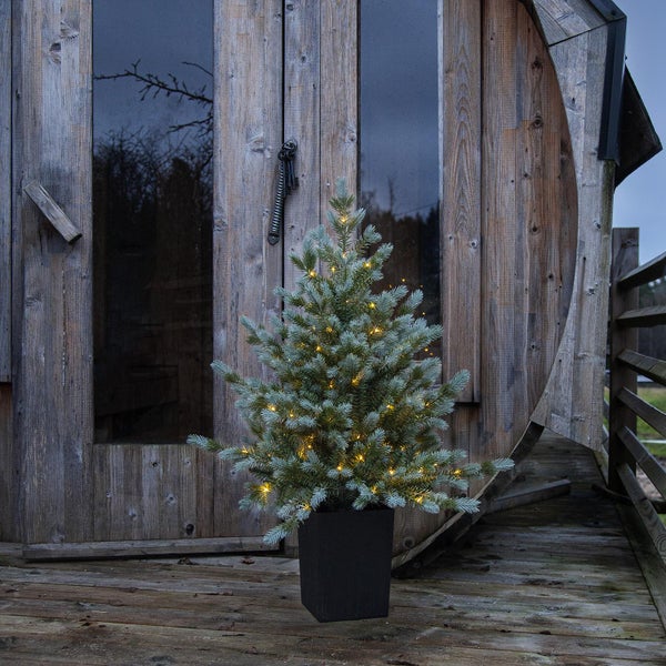 Künstlicher Tannenbaum mit Beleuchtung im Topf vor einem Holzhaus.
