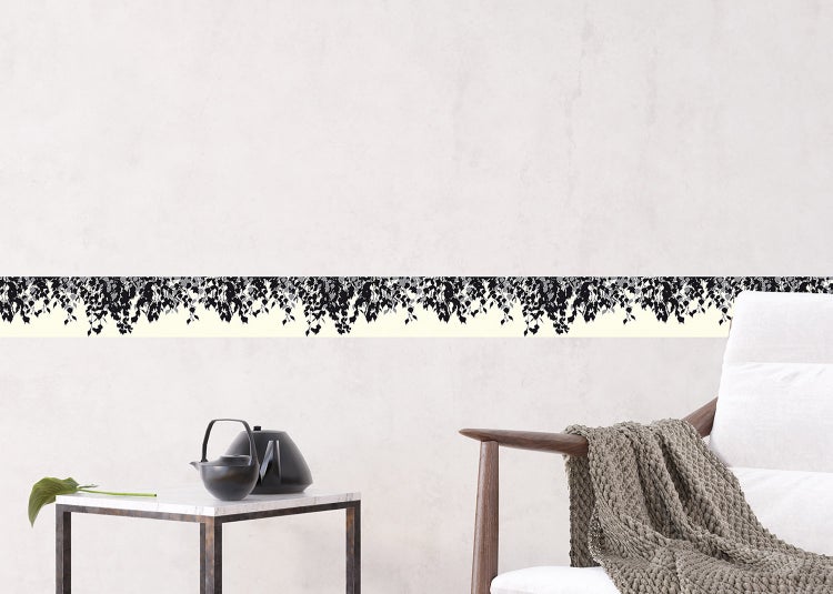 Selbstklebende Tapetenbordüre mit schwarzem Blättermuster an einer hellen Wand in einem Wohnraum mit Beistelltisch und Sessel.