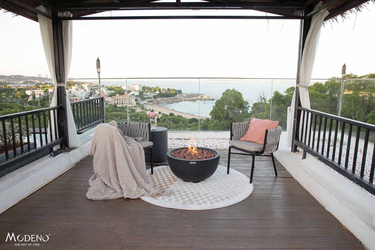 Helle Terrasse mit runder Modeno Gasfeuerschale, Loungesesseln, Holzdeck und Küstenpanorama. Modeno Logo.