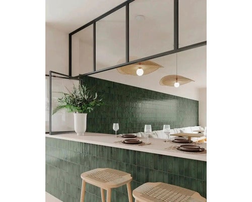 Moderne Bar mit dunkelgrünen Wandfliesen, hellem Tresen, Barhockern aus Holz und großen Sprossenfenstern mit schwarzem Rahmen.