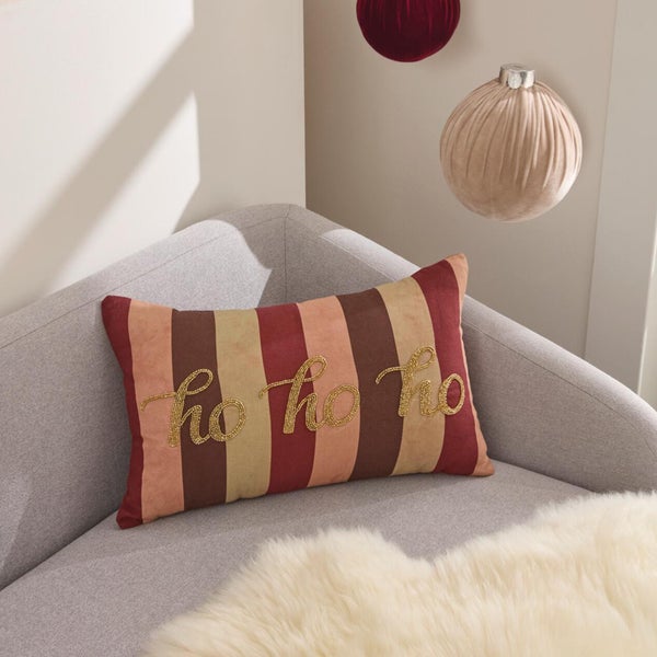 Weihnachtliches Dekokissen mit gestreiftem Muster und goldenem Ho ho ho Schriftzug auf einem Sofa mit Kunstfell.