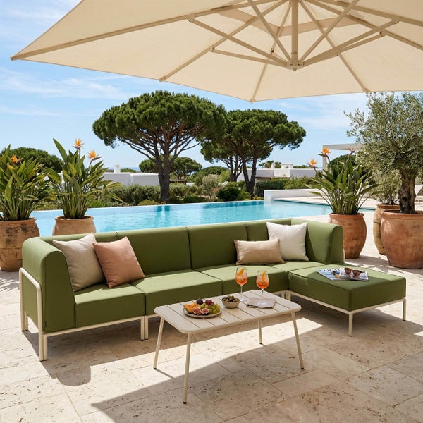 Moderne Gartenlounge mit grünem Ecksofa und Couchtisch auf einer hellen Terrasse mit Sonnenschirm und Pool im Hintergrund.
