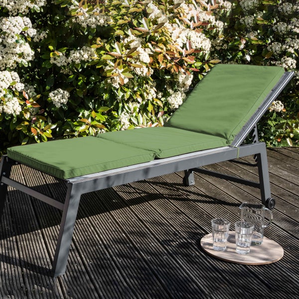 Gartenliege mit grüner Auflage auf einer Holzterrasse vor einer blühenden Hecke.