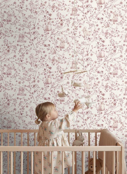 Kind in einem Kinderbett aus Holz vor einer Wand mit Tapete mit Hasen- und Blumenmuster, ein Mobile greifend.