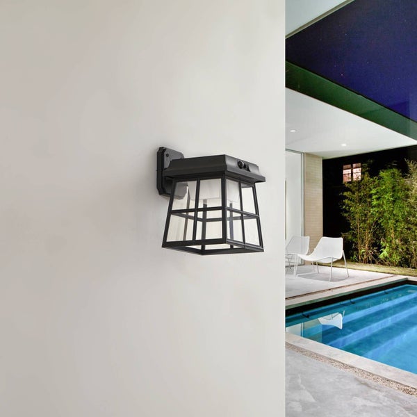 Schwarze Außenwandleuchte im Laternen-Stil mit Glaseinsätzen, montiert an einer hellen Fassade im Poolbereich.