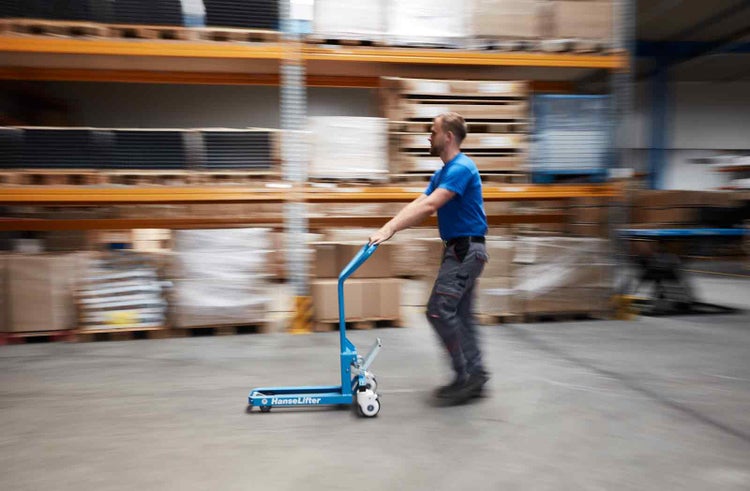 Ein Mann zieht einen blauen HanseLifter Hubwagen in einem Lagerhaus mit Regalen voller Waren.
