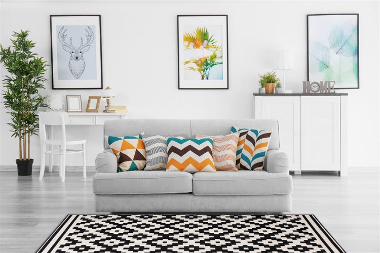 Helles Wohnzimmer mit Sofa, gemusterten Kissen, Beistelltisch mit Stuhl, Sideboard und geometrischem Teppich als moderne Wohndekoration.