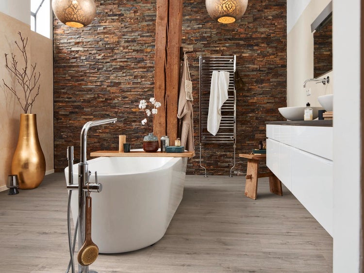 Modernes Badezimmer mit freistehender Badewanne, Wandverkleidung aus Naturstein, Holzbalken, Waschtisch und Holzoptik Bodenfliesen.