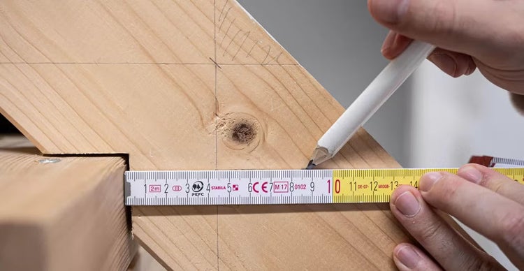 Eine Person markiert mit einem Bleistift und einem Gliedermaßstab der Marke Stabila eine Messung auf einem Holzbalken.