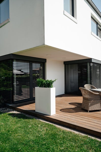 Hoher weißer Pflanzkübel mit grüner Pflanze auf einer Holzterrasse neben Korbstühlen vor einem modernen Haus mit dunklen Außenjalousien.