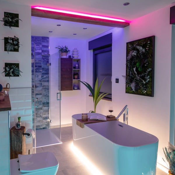 Modernes Badezimmer mit freistehender Badewanne, Glasdusche und stimmungsvoller LED-Beleuchtung in Pink und Blau. Wanddekoration mit Pflanzen.