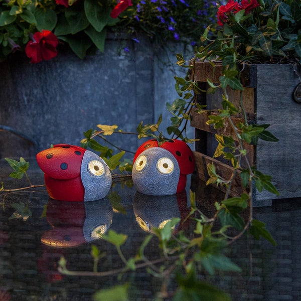 Zwei leuchtende Marienkäfer-Gartenstecker aus Stein und Kunststoff vor Pflanzen und Holzdeko