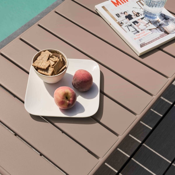 Gartentisch aus Metall mit Lamellenoberfläche, dekoriert mit einem Teller Pfirsiche, einer Schale Cracker, einer Zeitschrift und einem Glas Wasser.